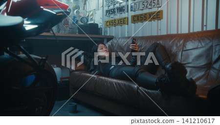 Female Lies Comfortably on Leather Couch Near Motorcycle, Enjoys Leisure Time in Garage Female Lies Comfortably on Leather Couch Near Motorcycle, Enjoys Leisure Time in Garage 141210617