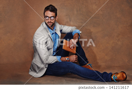 Fashion, book and man with glasses in studio with trendy, stylish and classy suit on floor. Reading, story and handsome male person with elegant style for outfit and novel by brown background. 141211617