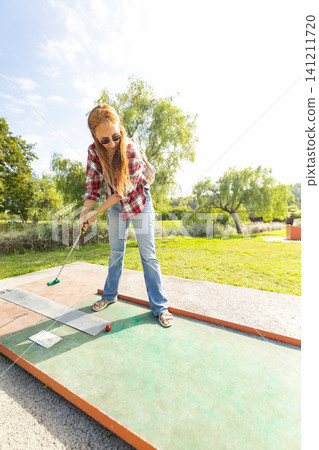 Girl plays mini golf in a sunny park while wearing sunglasses on a clear day in the afternoon during a fun outing with friends Girl plays mini golf in a sunny park while wearing sunglasses on a clear day in the afternoon during a fun outing with friends 141211720
