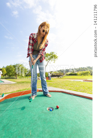 Woman plays mini golf on a sunny day at an outdoor course with green grass and trees in the background Woman plays mini golf on a sunny day at an outdoor course with green grass and trees in the background 141211796