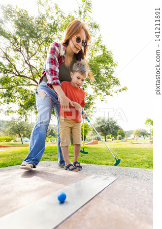 Mother and son play mini golf together at a park on a sunny day while practicing their swing and enjoying the outdoors Mother and son play mini golf together at a park on a sunny day while practicing their swing and enjoying the outdoors 141211891