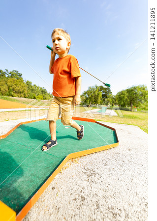 Child walks on mini golf course holding putter during sunny day outdoors Child walks on mini golf course holding putter during sunny day outdoors 141211895