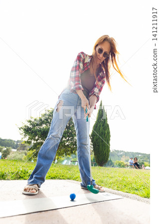 Woman plays mini golf on a sunny day in a park while wearing casual clothes and sunglasses 141211917