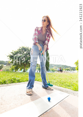 Woman plays mini golf in park during sunny afternoon with family in background Woman plays mini golf in park during sunny afternoon with family in background 141211923