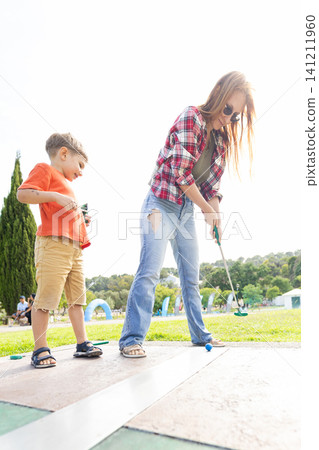 Woman and boy play mini golf together at a park on a sunny day with green trees in the background Woman and boy play mini golf together at a park on a sunny day with green trees in the background 141211960