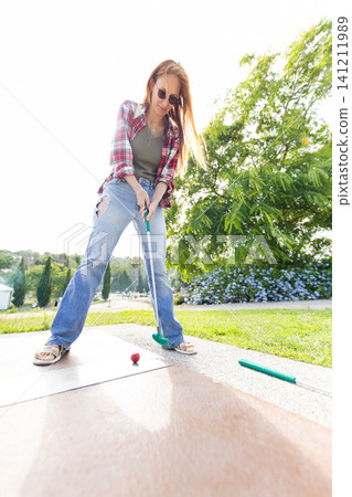 Woman practicing mini golf on a sunny day in a green park with trees and flowers around Woman practicing mini golf on a sunny day in a green park with trees and flowers around 141211989