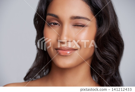 Woman, face and wink with beauty and skincare glow, playful and fun with lip gloss for moisturizer on grey background. Flirt in portrait, natural cosmetics and hair care with wellness and dermatology 141212433