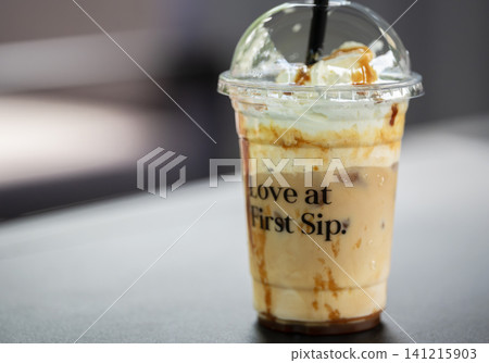 Angled shot of a chilled caramel coffee drink topped with cream and syrup in a plastic cup with a lid 141215903