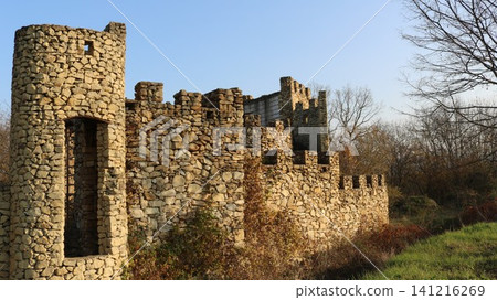 The abandoned ruins of a medieval stone castle with a round tower and crenellated walls. Sunlight illuminates the textured stonework against a backdrop of autumn trees and a clear sky. 141216269