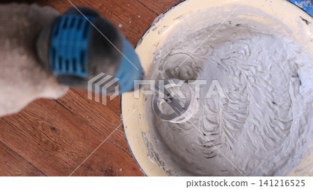 An overhead view of a worker mixing gray mortar in a yellow bowl. Using a drill and a special whisk attachment, the worker thoroughly mixes the thick putty on a wooden floor. An overhead view of a worker mixing gray mortar in a yellow bowl. Using a drill and a special whisk attachment, the worker thoroughly mixes the thick putty on a wooden floor. 141216525