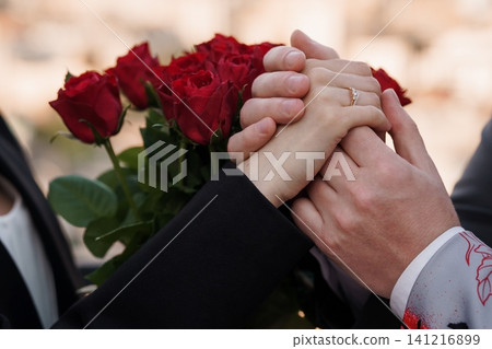 Close up of couple holding hands with engagement ring and red rose bouquet in formal black attire. Close up of couple holding hands with engagement ring and red rose bouquet in formal black attire. 141216899