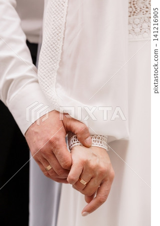 Close up of a couple holding hands showing gold wedding rings and white clothing in a soft romantic style. Close up of a couple holding hands showing gold wedding rings and white clothing in a soft romantic style. 141216905