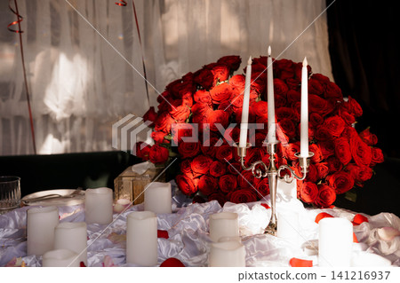 Romantic arrangement of red roses bouquet and lit white candles on a white fabric table with silver candelabra. 141216937