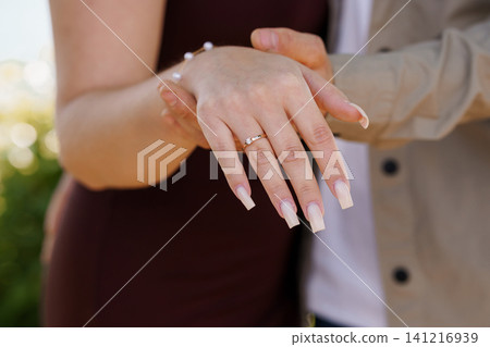 Close up of woman wearing diamond engagement ring and pearl bracelet held by man in beige jacket outdoors. Close up of woman wearing diamond engagement ring and pearl bracelet held by man in beige jacket outdoors. 141216939