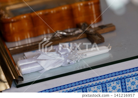 Decorated white wedding candles with ribbons and pearls resting on glass near a wooden crucifix Decorated white wedding candles with ribbons and pearls resting on glass near a wooden crucifix 141216957