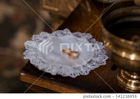 Two gold wedding rings resting on a white lace cushion on a dark wooden altar surface. Two gold wedding rings resting on a white lace cushion on a dark wooden altar surface. 141216958