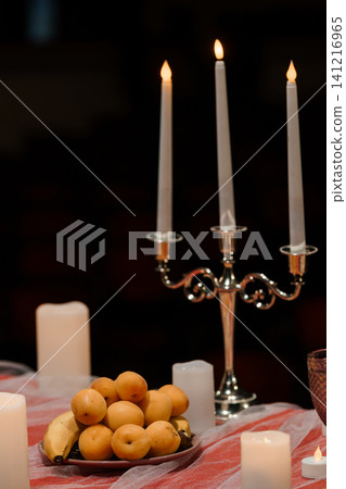 Lit white taper candles in silver candelabra with fresh yellow fruit on red tablecloth against dark background. 141216965