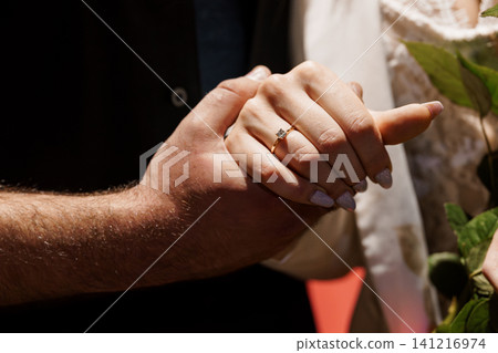 Close up of man and woman holding hands showing gold engagement ring with diamond on finger in soft sunlight. Close up of man and woman holding hands showing gold engagement ring with diamond on finger in soft sunlight. 141216974