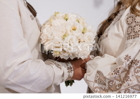 Couple in traditional embroidered white linen shirts holding a white flower bouquet against a plain background. Couple in traditional embroidered white linen shirts holding a white flower bouquet against a plain background. 141216977