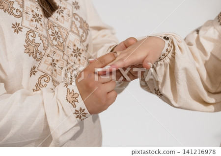 Person placing a ring on a finger wearing traditional embroidered linen clothing against a white background. Person placing a ring on a finger wearing traditional embroidered linen clothing against a white background. 141216978