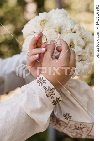 Close up of hands holding two gold wedding rings against white bridal flowers and embroidered linen clothing. Close up of hands holding two gold wedding rings against white bridal flowers and embroidered linen clothing. 141216983