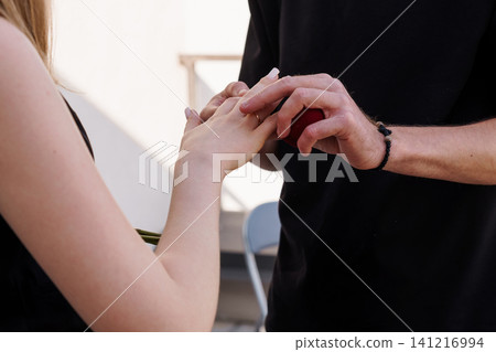 Man placing engagement ring on woman hand during romantic proposal in outdoor bright minimalist setting. Man placing engagement ring on woman hand during romantic proposal in outdoor bright minimalist setting. 141216994