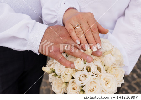 Close up of couple hands with wedding rings holding a white rose bouquet in traditional white clothing. Close up of couple hands with wedding rings holding a white rose bouquet in traditional white clothing. 141216997
