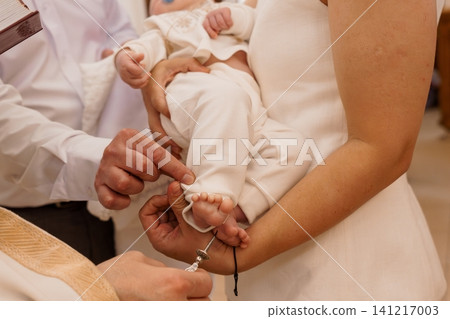 Priest anointing baby foot during baptism ceremony in church with white clothing and warm lighting. 141217003