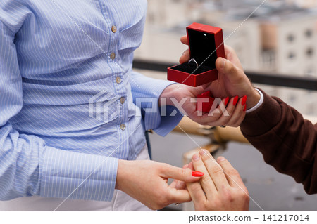 Man Proposing Marriage with Diamond Ring in Red Box to Woman on Urban Rooftop during Daytime. Man Proposing Marriage with Diamond Ring in Red Box to Woman on Urban Rooftop during Daytime. 141217014