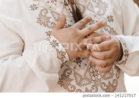 Man wearing traditional embroidered linen shirt touching gold wedding ring on finger in soft natural light. 141217017