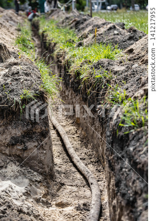 Workers are laying a high voltage cable in a trench at a construction site, with dirt and grass along the edges and bright daylight present Workers are laying a high voltage cable in a trench at a construction site, with dirt and grass along the edges and bright daylight present 141217955