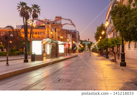 Evening view of Avenida de la Constitucion in Seville, Spain, with illuminated tram stop, historic architecture, and empty pedestrian boulevard in the city center. Evening view of Avenida de la Constitucion in Seville, Spain, with illuminated tram stop, historic architecture, and empty pedestrian boulevard in the city center. 141218033
