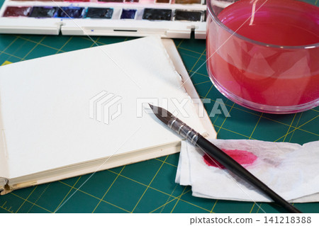 Paintbrush on napkin by blank sketchpad, glass jar filled with pink tinted water on green cutting mat, creative art workspace Paintbrush on napkin by blank sketchpad, glass jar filled with pink tinted water on green cutting mat, creative art workspace 141218388