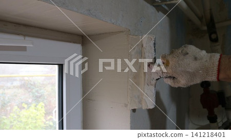 A close-up of a hand in a work glove applying mortar to a window reveal with a spatula. The process of leveling corners and walls takes place indoors during renovations. A close-up of a hand in a work glove applying mortar to a window reveal with a spatula. The process of leveling corners and walls takes place indoors during renovations. 141218401