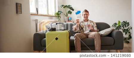 Image of handsome male model, tourist with suitcase, shows his two tickets on plane, uses mobile phone app, sits on sofa Image of handsome male model, tourist with suitcase, shows his two tickets on plane, uses mobile phone app, sits on sofa 141219080