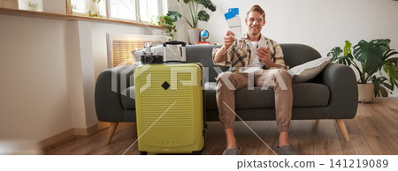 Portrait of happy man checked in online on smartphone, showing tickets in his hand, sitting near suitcase, going on vacation, preparing for holiday 141219089