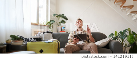 Young man hoping for something, tourist with digital tablet, cross fingers, sits with suitcase packed, praying for holiday Young man hoping for something, tourist with digital tablet, cross fingers, sits with suitcase packed, praying for holiday 141219115