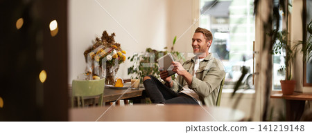 Portrait of happy, relaxed young man sitting in cafe alone, wearing wireless headphones, playing video game on tablet, laughing and smiling, looking at his device. Concept of lifestyle and people Portrait of happy, relaxed young man sitting in cafe alone, wearing wireless headphones, playing video game on tablet, laughing and smiling, looking at his device. Concept of lifestyle and people 141219148