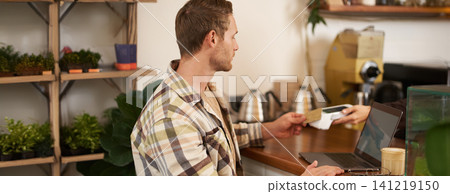 Portrait of freelancer, young man with laptop in coffee shop, paying for his order, pressing his credit card to the POS terminal, contactless payment concept Portrait of freelancer, young man with laptop in coffee shop, paying for his order, pressing his credit card to the POS terminal, contactless payment concept 141219150