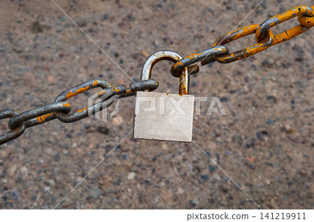 An old metal chain with peeling orange paint, connected by a hanging steel lock against the background of stony or gravel old asphalt. A symbol of closed access, the end of a free path 141219911