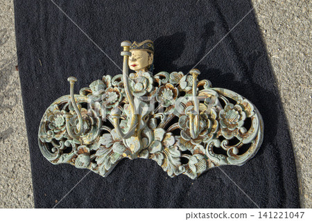 XALO, SPAIN - JUNE 20, 2026.A vintage wall coat hanger shows detailed metal carvings in floral shapes with a hand painted Balinese face at the top. The hanger is on a black surface. XALO, SPAIN - JUNE 20, 2026.A vintage wall coat hanger shows detailed metal carvings in floral shapes with a hand painted Balinese face at the top. The hanger is on a black surface. 141221047