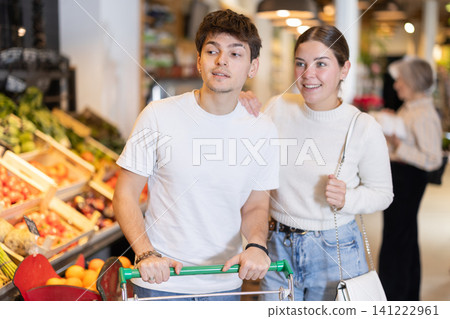 In food store, young woman and guy with trolley looking for something on shelf of showcase In food store, young woman and guy with trolley looking for something on shelf of showcase 141222961