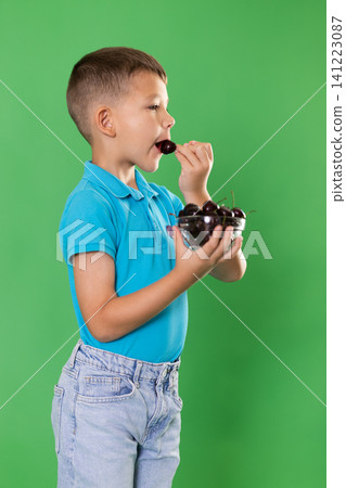 Boy in blue clothes happily eats ripe cherries on green background Boy in blue clothes happily eats ripe cherries on green background 141223087