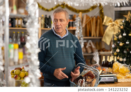 Mature male shopper with grocery basket attentively choosing fresh food and alcohol in grocery supermarket for Christmas celebration Mature male shopper with grocery basket attentively choosing fresh food and alcohol in grocery supermarket for Christmas celebration 141223232