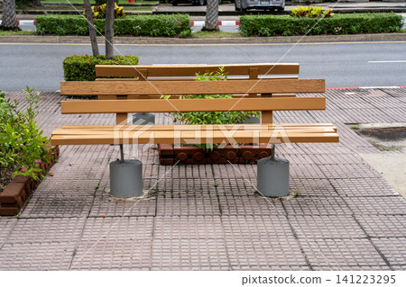 Modern wooden bench on a paved sidewalk with concrete bases. Public seating area in an urban park or street setting with green plants and road in the background. Professional outdoor furniture. 141223295