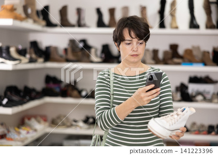 Female customer examines shoes in store and scans label with phone camera. Female customer examines shoes in store and scans label with phone camera. 141223396