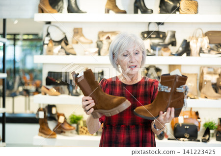 Elderly woman buyer looks at footwear from new collection and picks up new pair of boots for winter. Elderly woman buyer looks at footwear from new collection and picks up new pair of boots for winter. 141223425