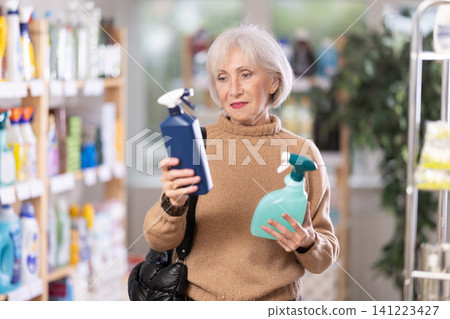 Elderly female shopper carefully selects a spray glass cleaner for windows in home Elderly female shopper carefully selects a spray glass cleaner for windows in home 141223427