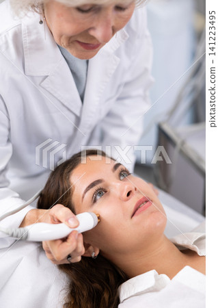 Mature female dermatologist performing cosmetology procedure for young woman, RF lifting and vacuum massage for facial skin therapy in clinic Mature female dermatologist performing cosmetology procedure for young woman, RF lifting and vacuum massage for facial skin therapy in clinic 141223495