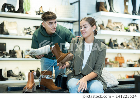 Young guy and girl choosing demi-season womens boots in shoe store Young guy and girl choosing demi-season womens boots in shoe store 141223524